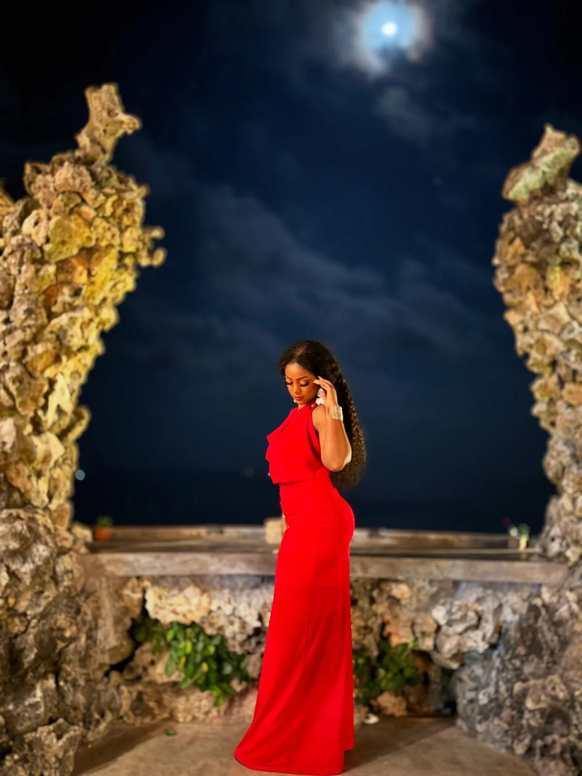 Indy, founder of Beauty by Indy, in a red evening gown beneath a full moon on a rocky oceanside terrace