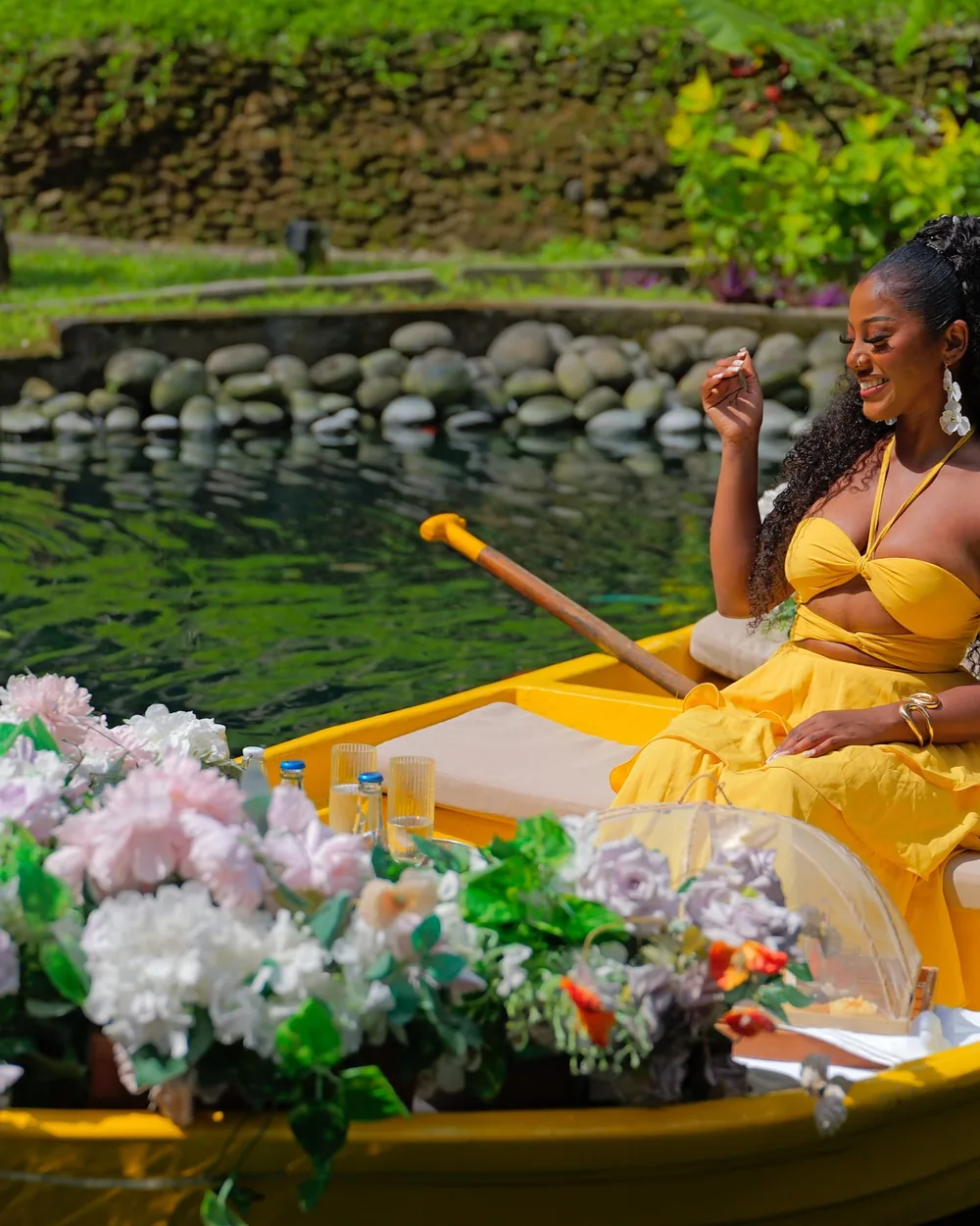 Indy on a flower-lined boat in a yellow dress, smiling in a tropical garden.